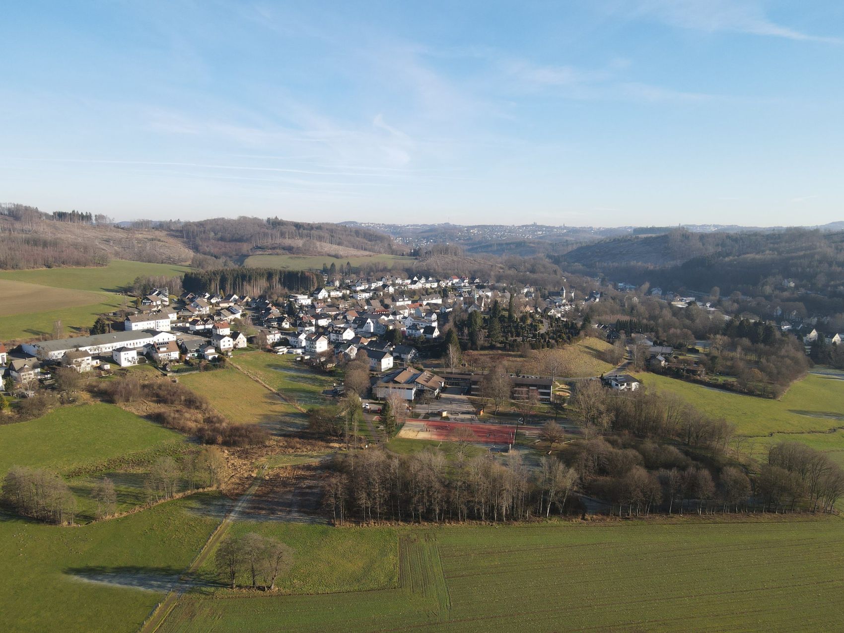 Luftaufnahme von Oberbrügge mit der Regenbogenschule im Zentrum, umgeben von Feldern und Wäldern.
