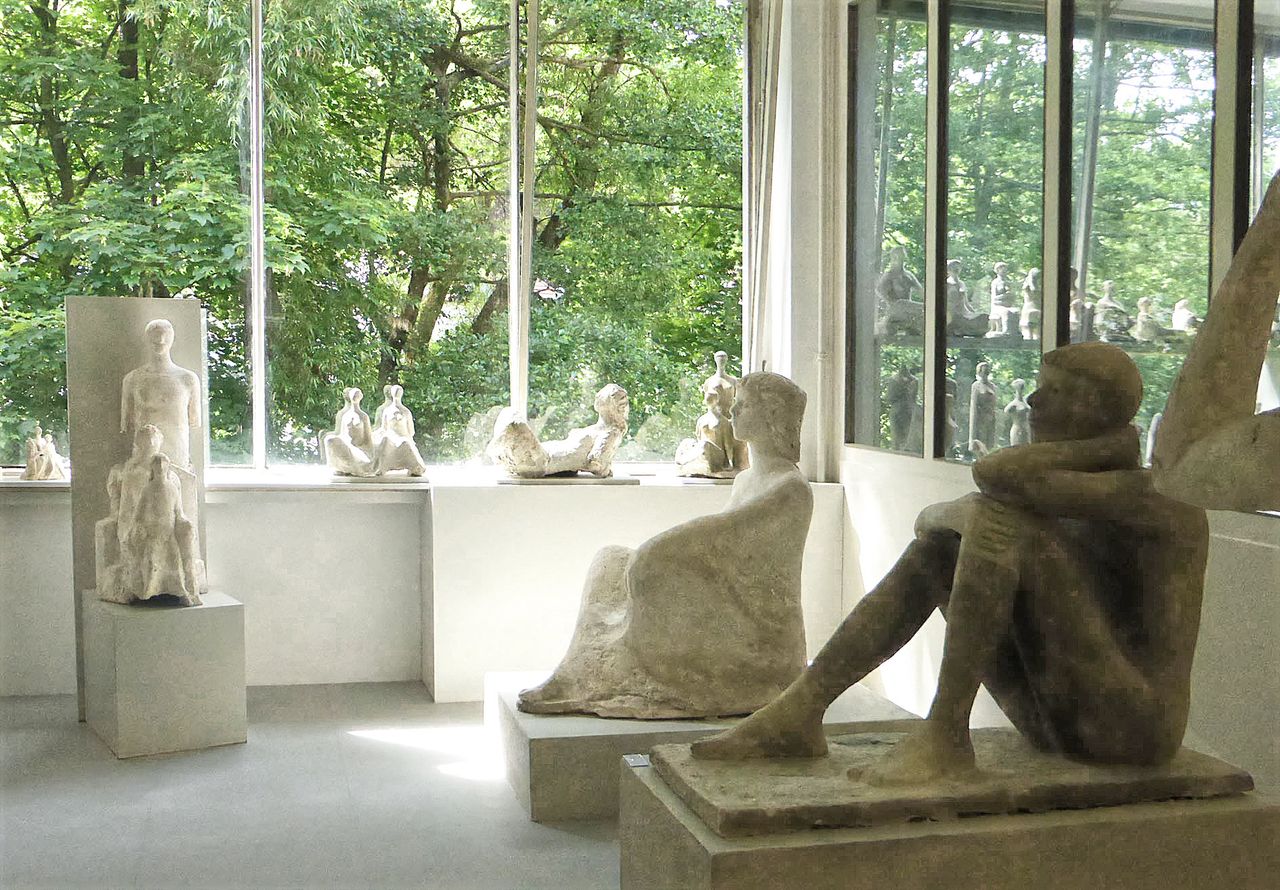 Ausstellungsraum mit mehreren Skulpturen aus hellem Stein, vor großen Fenstern mit Blick auf Grünflächen.