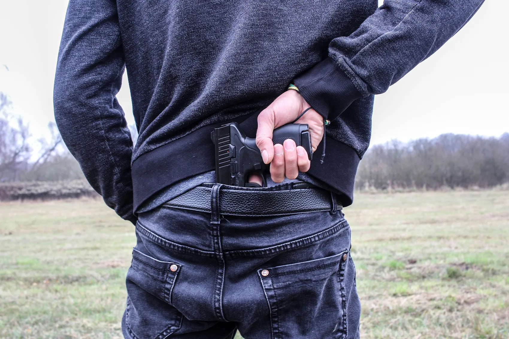 Eine Person hält eine Pistole im hinteren Hosenbund, vermutlich zur Selbstverteidigung. Im Hintergrund eine verschwommene Landschaft.