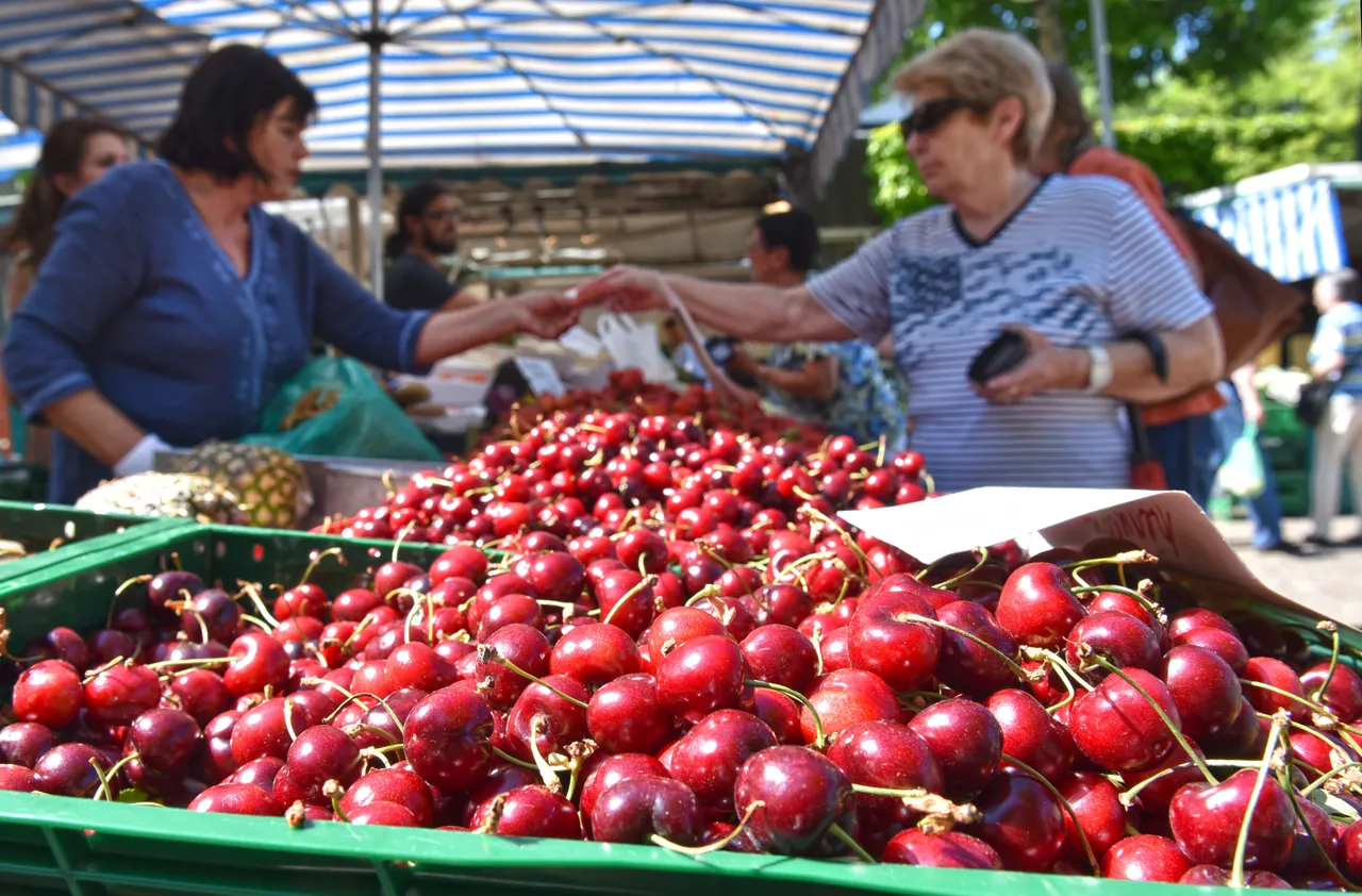 Ein Korb voller roter Kirschen im Vordergrund, dahinter zwei Frauen an einem Obststand unter einem blauen Sonnenschirm.