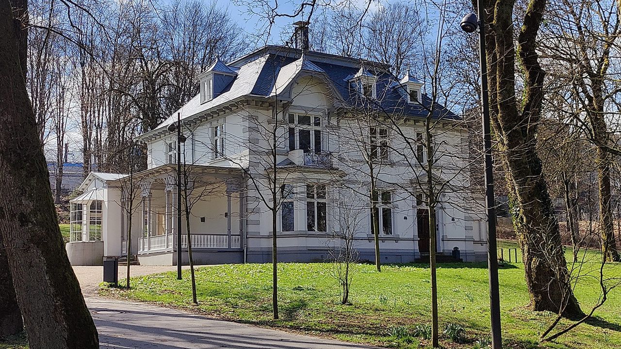 Helles, mehrgeschossiges Gebäude mit mehreren Fenstern und einem dunklen Dach, umgeben von Rasenflächen und Bäumen im Park von Meinerzhagen.