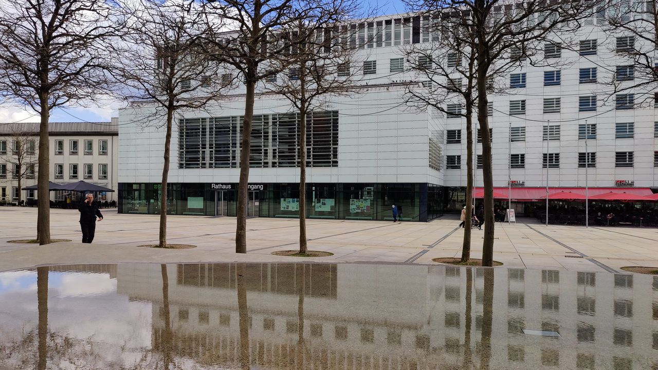 Moderner Verwaltungsbau mit Glasfassade und dem Eingangsbereich des Rathauses Lüdenscheid. Im Vordergrund spiegeln sich die Gebäude in einer nassen Pflasterung.