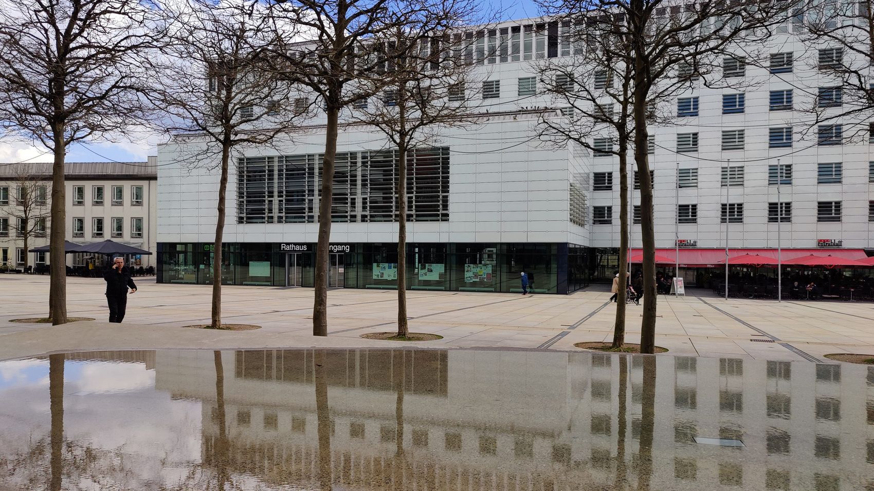 Moderner Verwaltungsbau mit Glasfassade und dem Eingangsbereich des Rathauses Lüdenscheid. Im Vordergrund spiegeln sich die Gebäude in einer nassen Pflasterung.