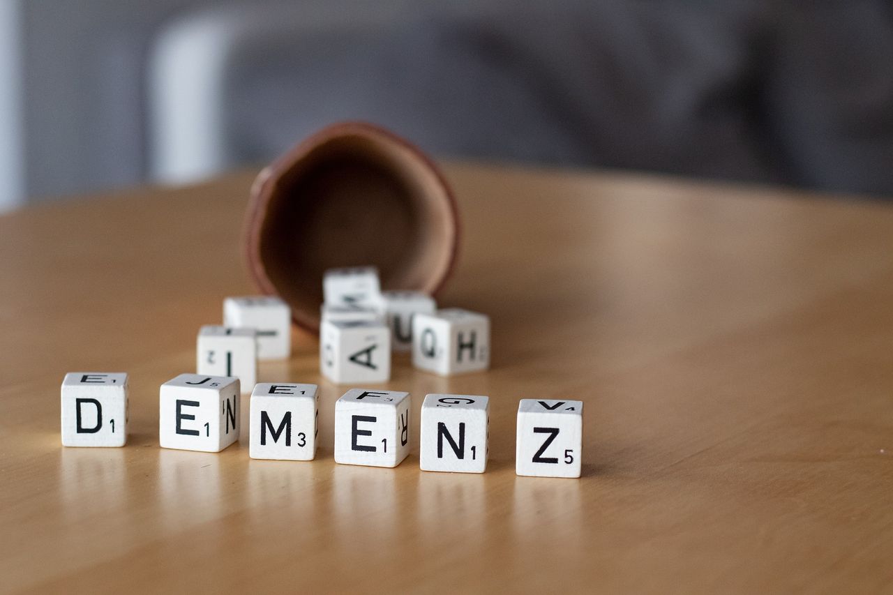 Weiße Buchstabensteine auf einem Holztisch bilden das Wort „DEMENZ“. Ein brauner Behälter liegt umgekippt im Hintergrund.