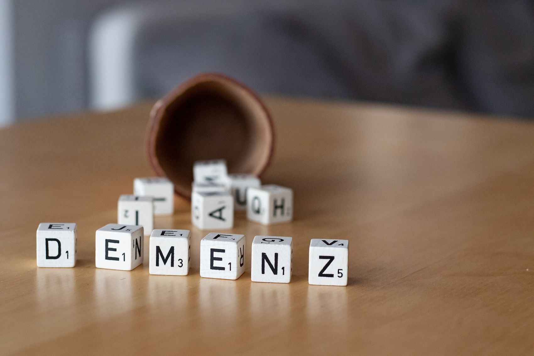Weiße Buchstabensteine auf einem Holztisch bilden das Wort „DEMENZ“. Ein brauner Behälter liegt umgekippt im Hintergrund.