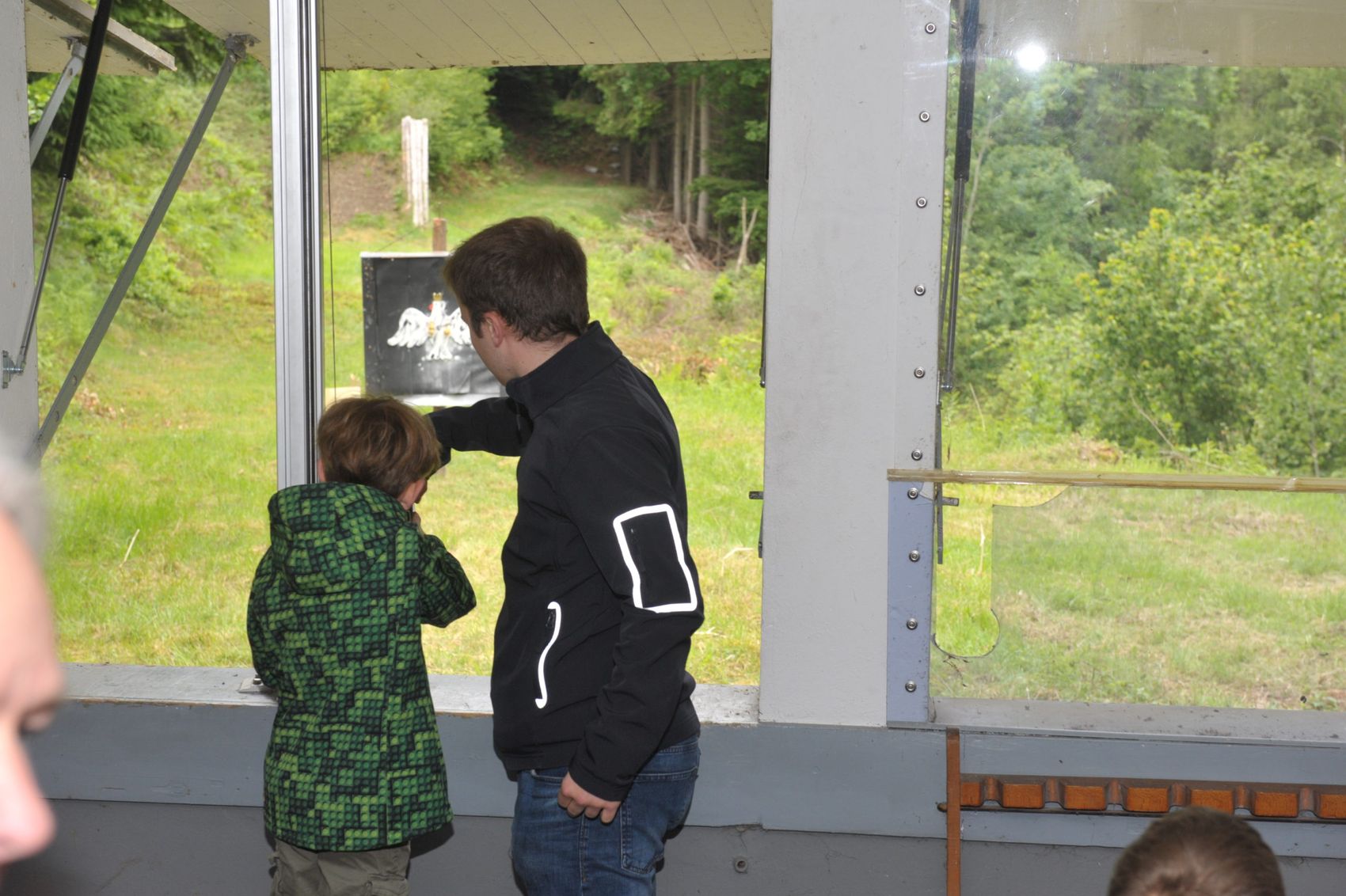 Ein Mann und ein Kind zielen mit Gewehren auf eine Zielscheibe im Freien, während sie sich in einem Schießstand befinden.