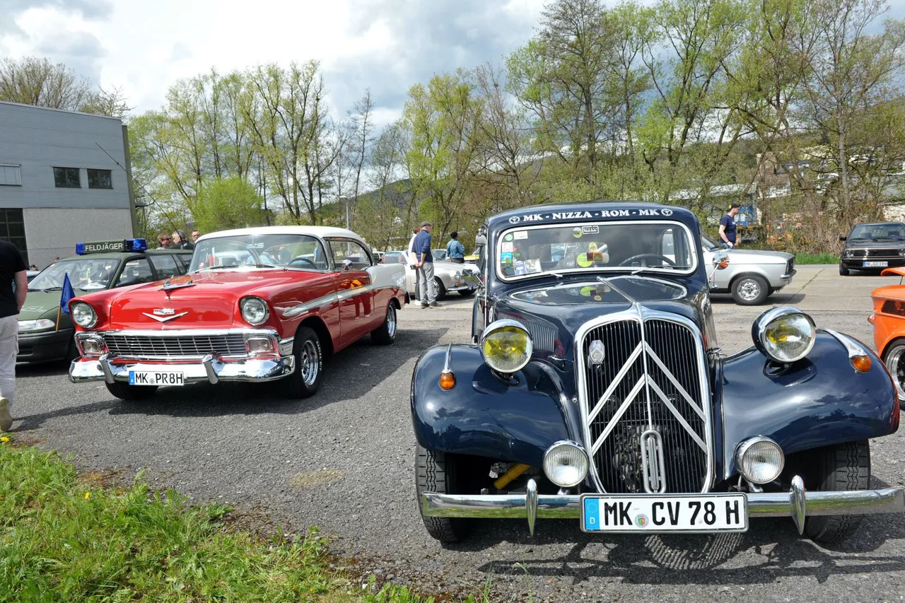 Dunkelblauer Citroën Traction Avant steht im Vordergrund eines Oldtimer-Treffens mit einem roten US-amerikanischen Oldtimer im Hintergrund.