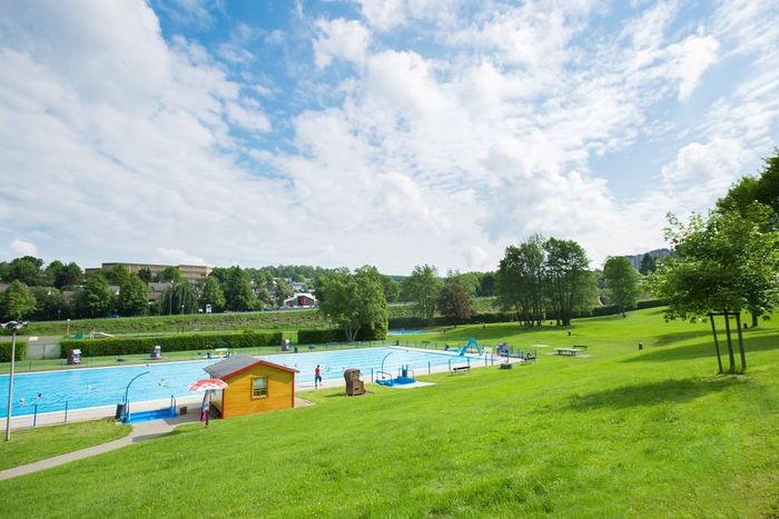 Ein sonniger Blick auf das Freibad Meinerzhagen mit Schwimmbecken, Liegewiese und einem kleinen orangefarbenen Gebäude. Bäume und Gebäude sind im Hintergrund zu sehen.