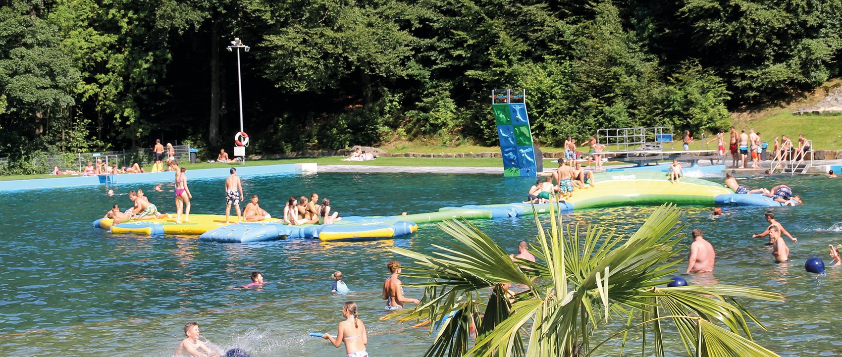 Menschen schwimmen und spielen auf einem aufblasbaren Wasserspielplatz in einem See, umgeben von Bäumen und einer Wiese.