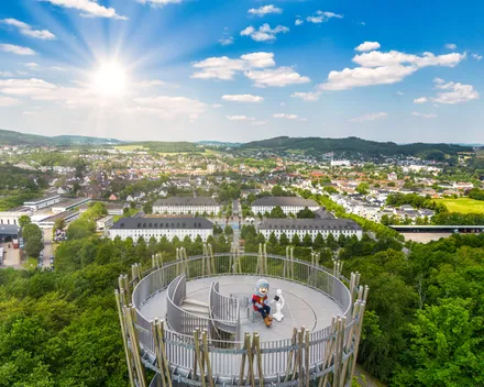 Ein Aussichtsturm mit Holzgeländer steht im Vordergrund, auf dem zwei Personen sitzen. Im Hintergrund erstreckt sich eine Stadtlandschaft mit Gebäuden und grünen Hügeln unter einem blauen Himmel mit Wolken.