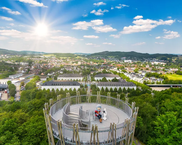 Ein Aussichtsturm mit Holzgeländer steht im Vordergrund, auf dem zwei Personen sitzen. Im Hintergrund erstreckt sich eine Stadtlandschaft mit Gebäuden und grünen Hügeln unter einem blauen Himmel mit Wolken.