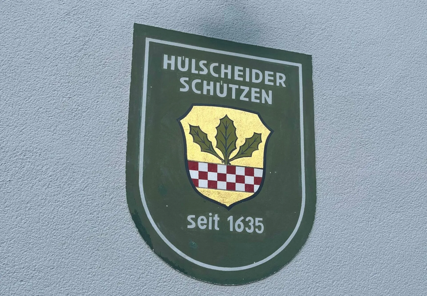 Grünes Schild mit der Aufschrift „HÜLSCHEIDER SCHÜTZEN“ und „seit 1635“ sowie einem Wappen mit einem Blatt und einem rot-weißen Schachbrettmuster.