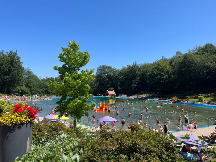 Viele Menschen planschen und entspannen an einem sonnigen Tag im Freibad Herpine, während sich Besucher am Beckenrand und unter Sonnenschirmen aufhalten.