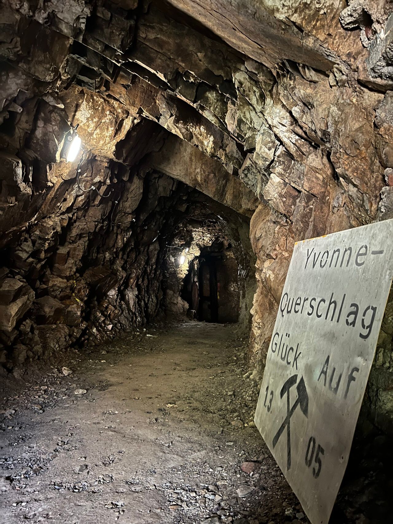 Ein dunkler Stollen in der Grube Neu Glück mit grobem Gestein an Wänden und Decke. Ein Schild mit der Aufschrift Yvonne-Querschlag, Glück Auf und weiteren Details steht im Vordergrund.