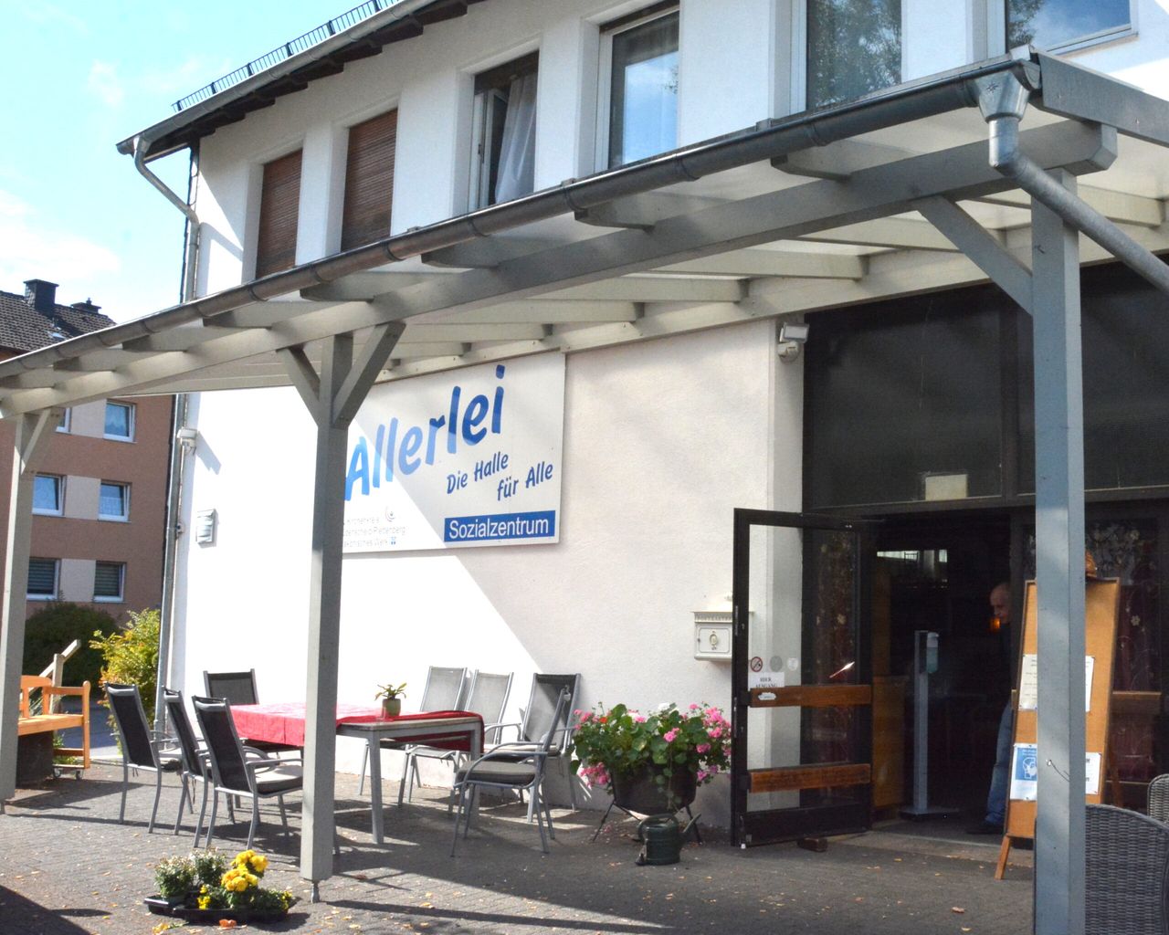 Außenansicht des Sozialzentrums Allerlei mit dem blauen Schriftzug an der weißen Fassade und einem überdachten Sitzbereich.