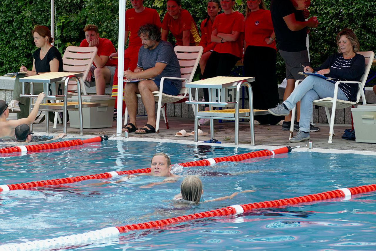 Zwei Schwimmer im Wasser, während Zuschauer auf Liegestühlen und Stühlen am Beckenrand sitzen. Im Hintergrund sind mehrere Personen in roten DLRG-Shirts zu sehen.