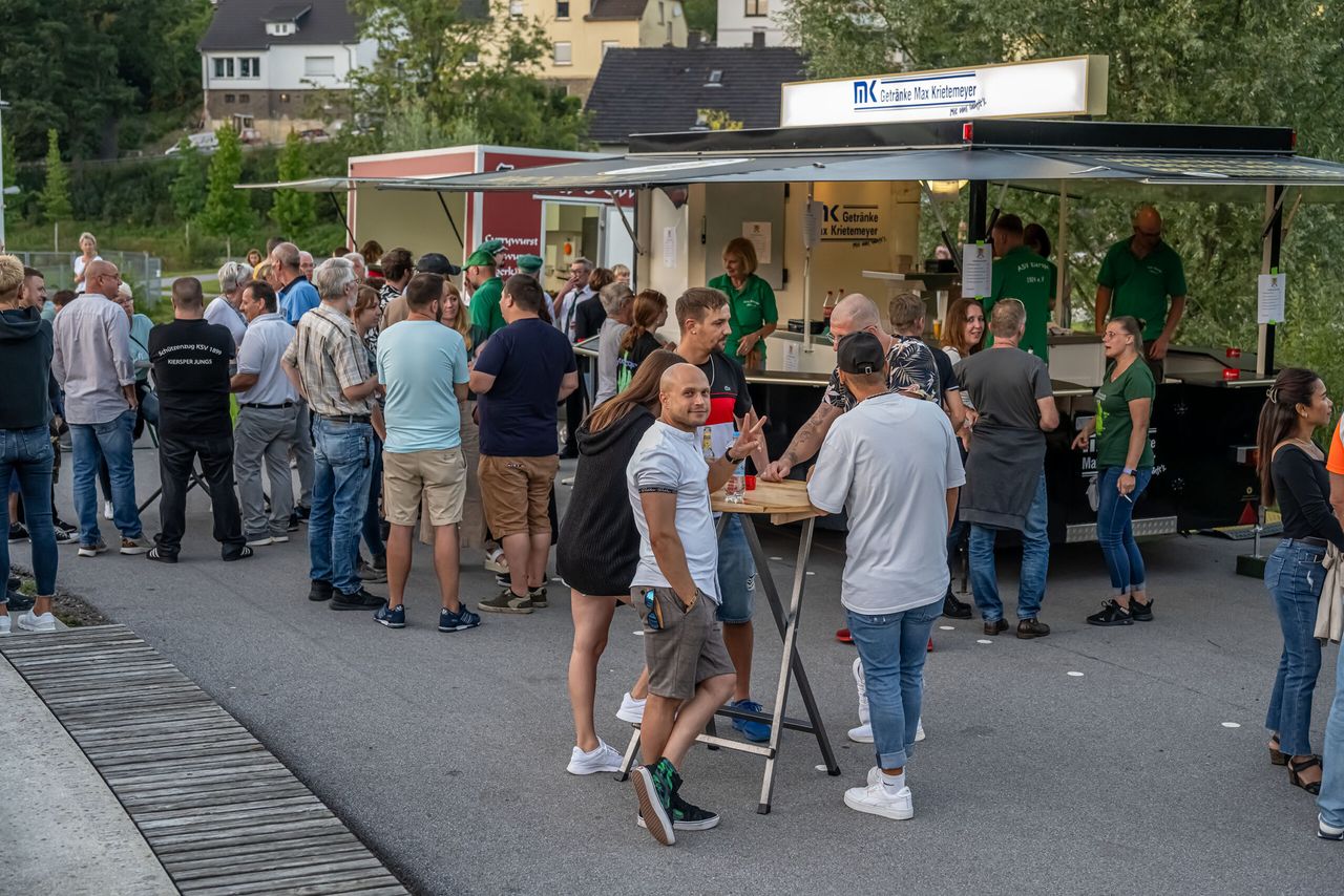 Menschen stehen und unterhalten sich vor mehreren Essensständen im Freien, während im Hintergrund ein DJ auflegt.