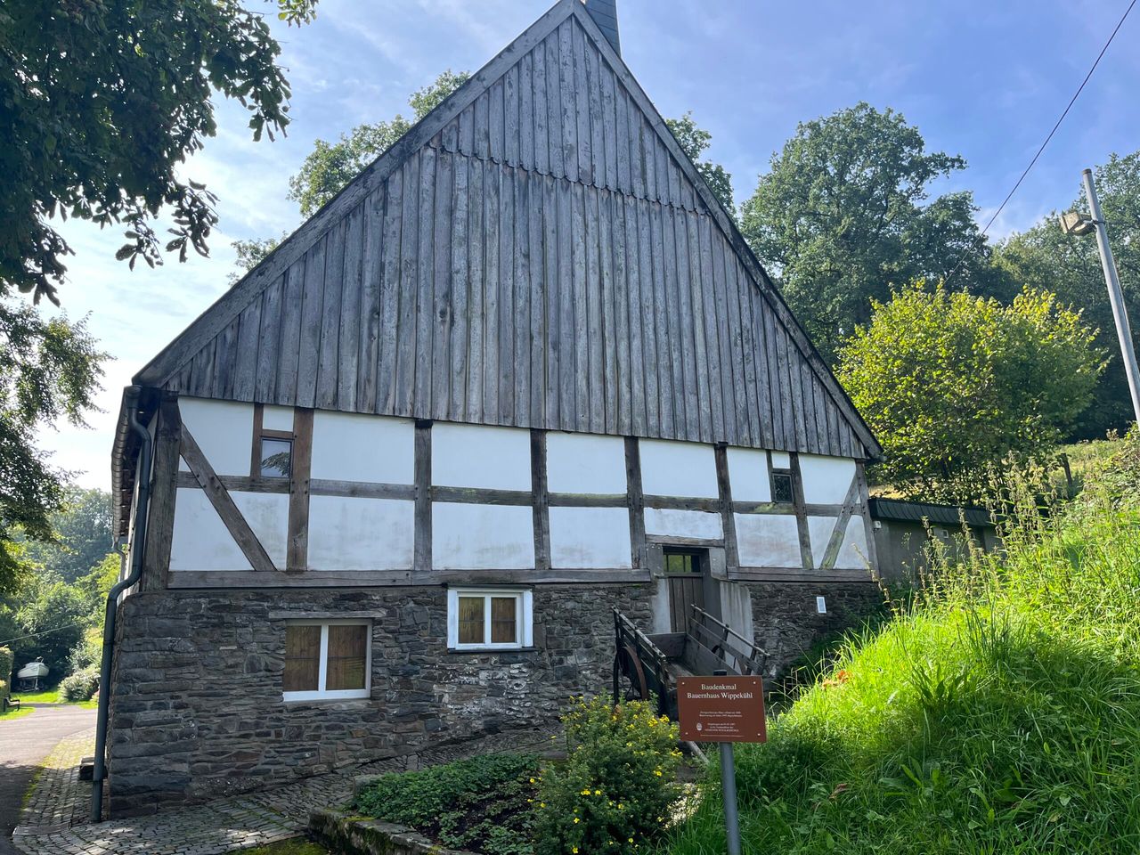 Fachwerkhaus mit grauem Schieferdach und Sockel aus Naturstein, davor eine Informationstafel.