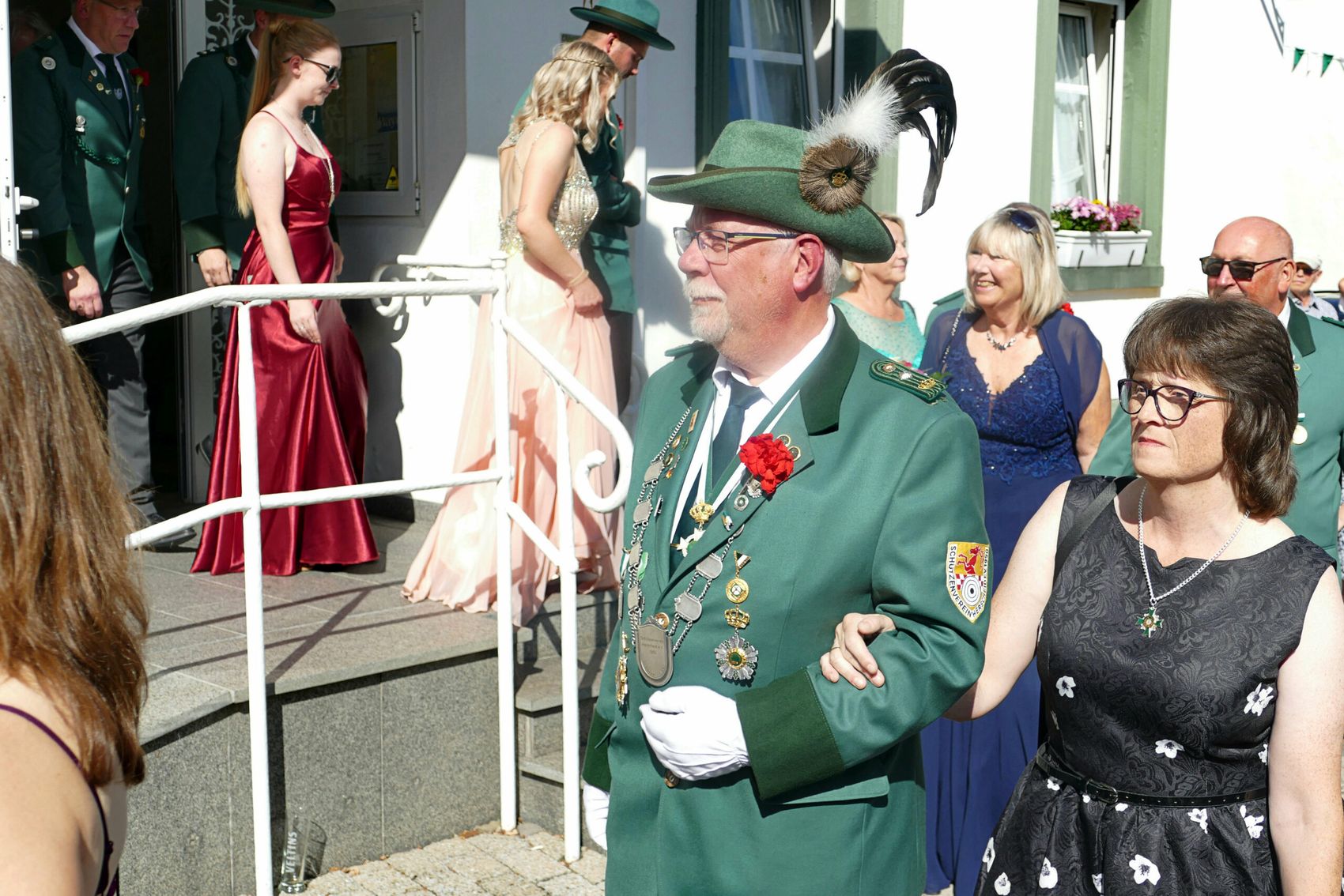Ein Mann in einem grünen Schützenumhang mit zahlreichen Orden und einer Feder auf dem Hut steht im Vordergrund. Im Hintergrund sind weitere Schützen in grünen Uniformen und festlich gekleidete Frauen zu sehen.