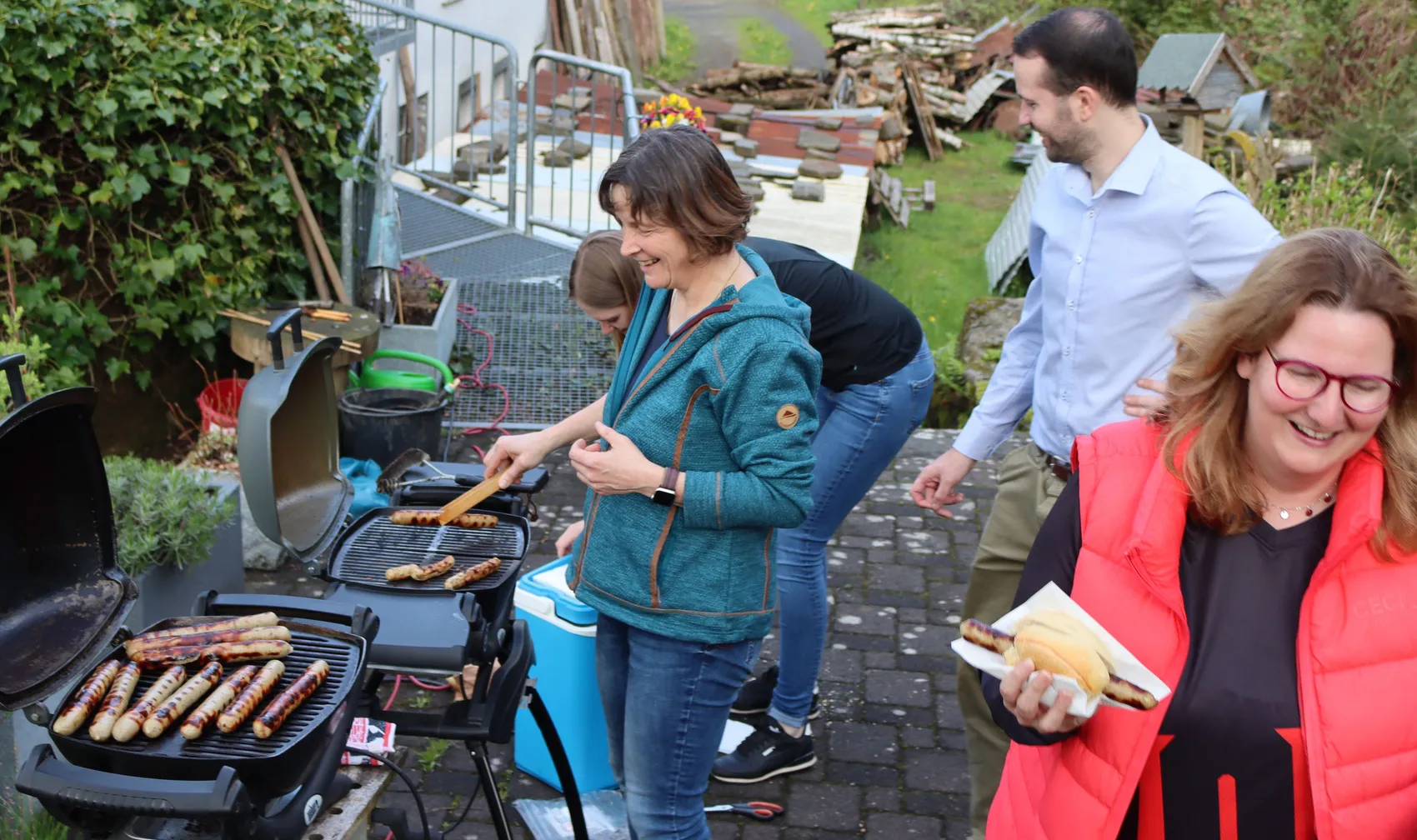 Eine Frau grillt Würstchen, während eine andere Person mit einer Wurst im Vordergrund lächelt. Im Hintergrund stehen weitere Personen und ein Grill.