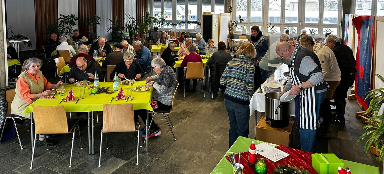 Menschen unterschiedlichen Alters sitzen an Tischen mit gelben Decken und nehmen in einem Gemeinschaftszentrum eine gemeinsame Mahlzeit ein.