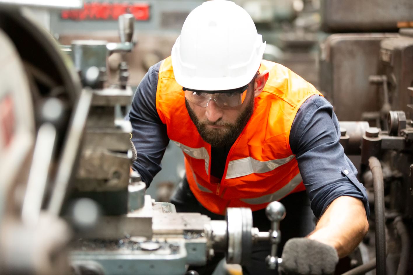 Ein Mann mit Bart, Schutzbrille und orangefarbener Warnweste bedient eine metallverarbeitende Maschine in einer Werkstatt.