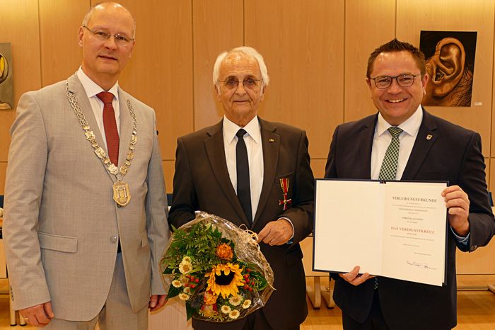 Klaus Ising steht mit einem Blumenstrauß und dem Bundesverdienstkreuz zwischen Ulrich Schulte und Marco Voge, der eine Urkunde hält.