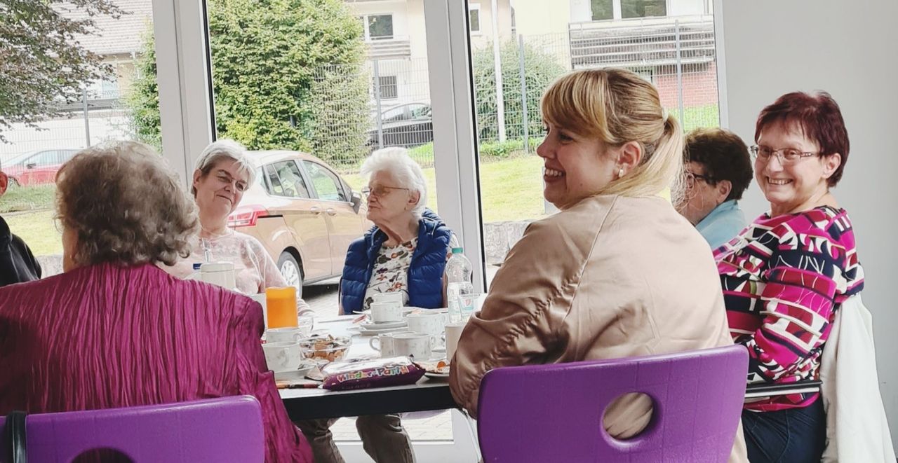 Eine Gruppe von Menschen sitzt an einem Tisch in einem hellen Raum und unterhält sich bei Getränken und Gebäck. Im Hintergrund ist durch ein Fenster ein parkender Wagen zu sehen.
