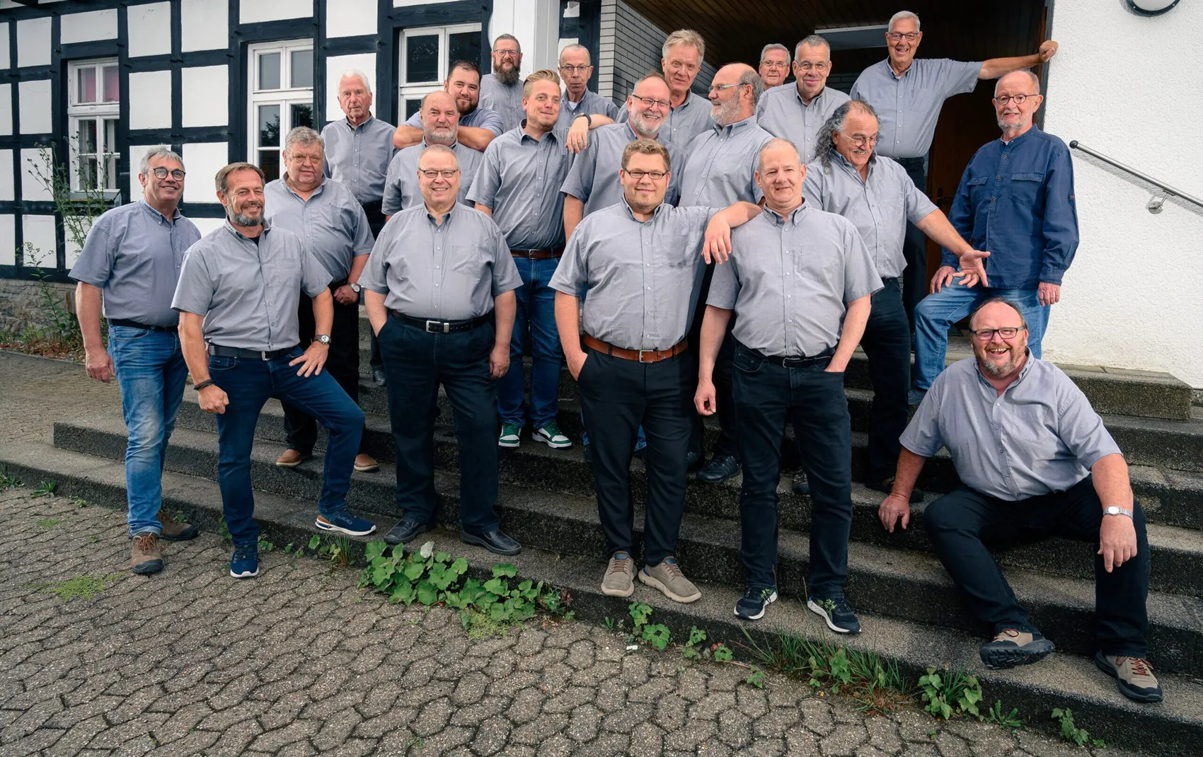 Eine Gruppe von Männern in hellgrauen Hemden posiert auf Treppenstufen vor einem Fachwerkhaus.