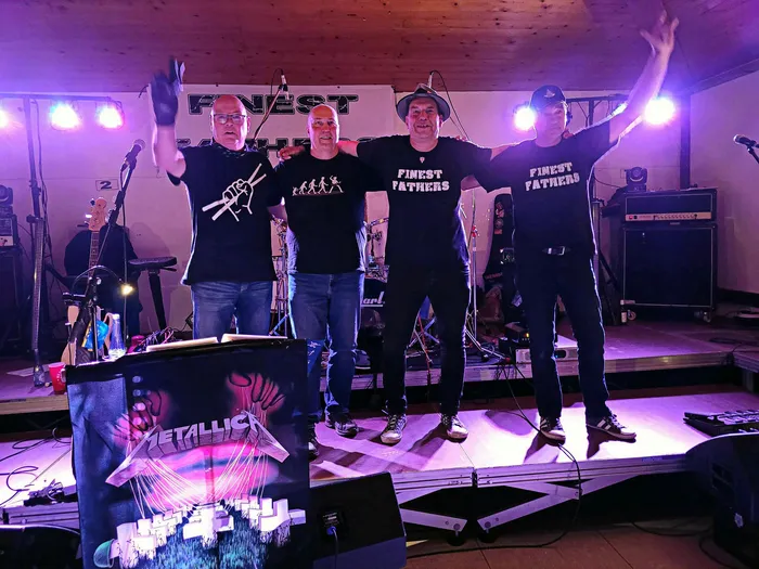 Vier Musiker stehen auf einer Bühne vor Schlagzeug und Verstärkern. Sie tragen T-Shirts mit Aufdruck und heben die Arme, während im Hintergrund ein Banner mit der Aufschrift „Finest“ zu sehen ist.