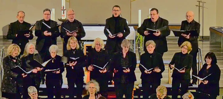 Chor singt in dunkler Kleidung mit Notenblättern in einer Kirche, im Hintergrund ein Altar und Kerzen.