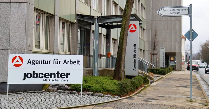 Das Bild zeigt das Gebäude der Agentur für Arbeit und des Jobcenters Märkischer Kreis. Im Vordergrund steht ein Schild mit dem Logo der Agentur für Arbeit und dem Hinweis