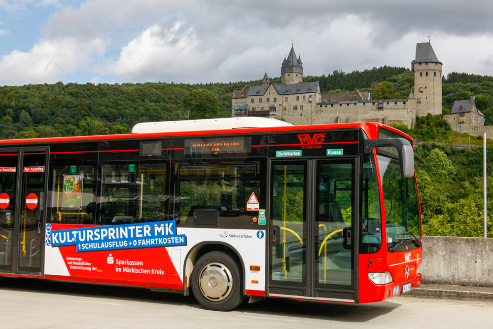 Roter Bus mit Aufschrift „Kultursprinter MK – 1 Schulausflug + 0 Fahrtkosten“ vor einer mittelalterlichen Burg auf einem bewaldeten Hügel.