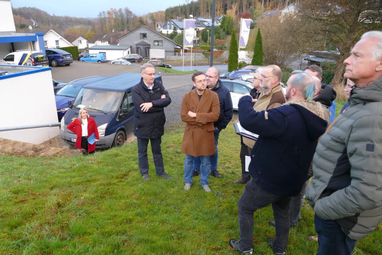 Mehrere Personen stehen auf einer Wiese und besprechen Pläne, während im Hintergrund ein Parkplatz mit Autos und Häusern zu sehen ist.