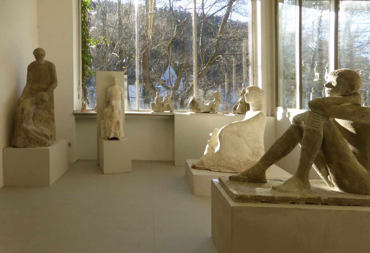 Raum mit mehreren grossformatigen Skulpturen aus hellem Stein, die auf Sockeln stehen. Im Hintergrund ist durch grosse Fenster ein Wald zu sehen.