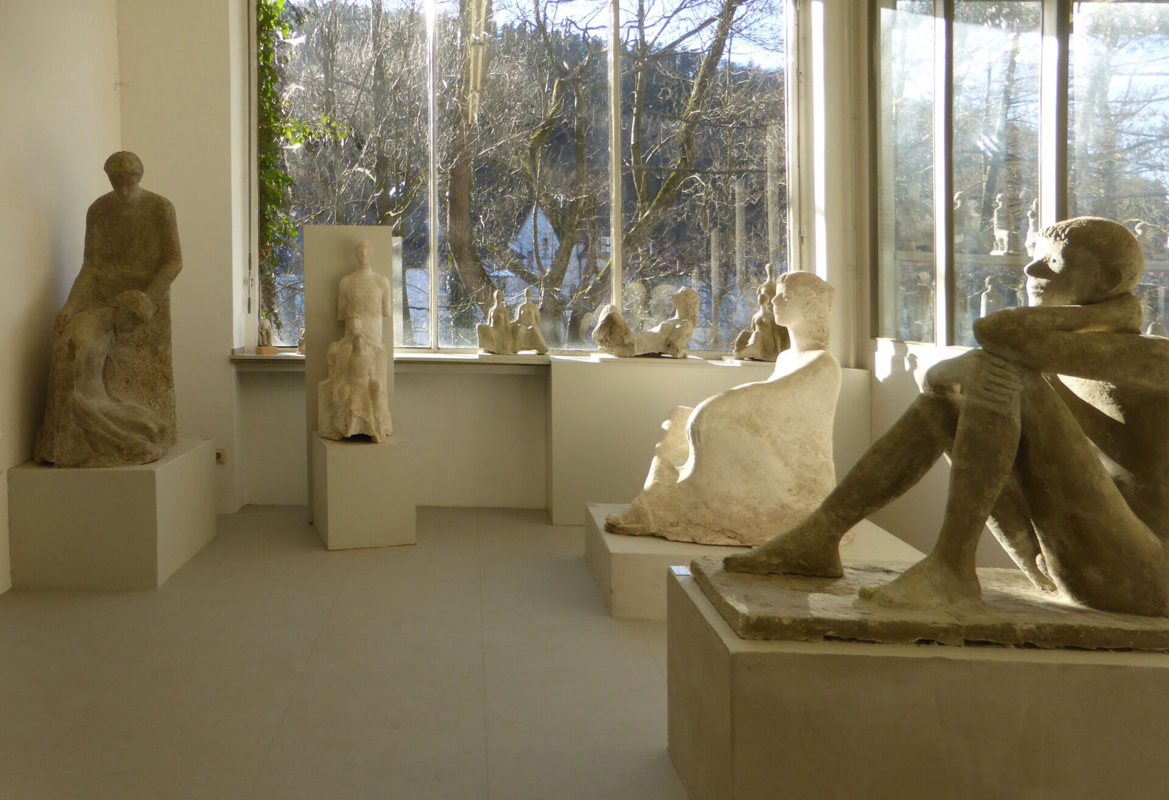 Raum mit mehreren grossformatigen Skulpturen aus hellem Stein, die auf Sockeln stehen. Im Hintergrund ist durch grosse Fenster ein Wald zu sehen.