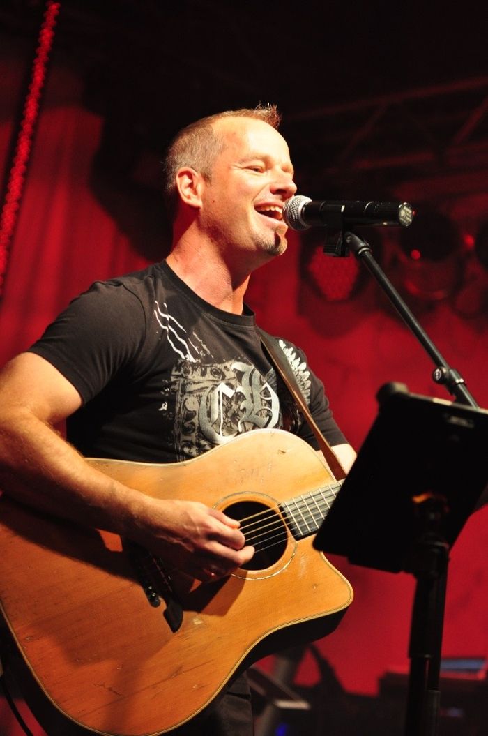 George Schneider spielt Akustikgitarre und singt in ein Mikrofon auf einer Bühne.