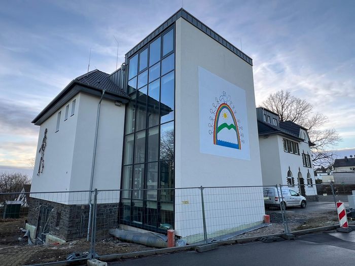Die Grundschule Wiblingwerde mit einer großen Glasfassade, einem bunten Regenbogen-Wandbild und einem Bauzaun im Vordergrund.