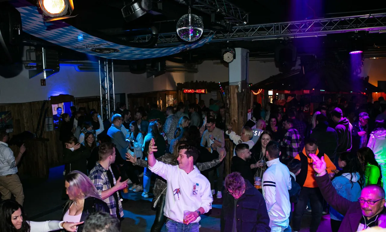 Tanzende Menschen feiern in einer festlich beleuchteten Halle mit Holztäfelung und einer Spiegeldiskokugel an der Decke.