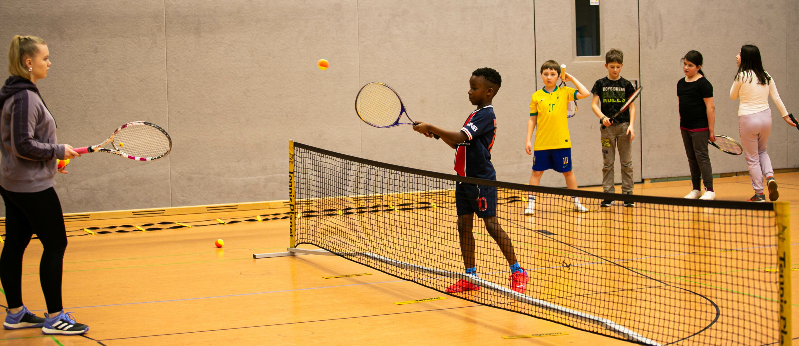 Zwei Kinder spielen Tischtennis in einer Turnhalle, während weitere Kinder zusehen. Ein Junge mit dunkler Hautfarbe schlägt den Ball über das Netz.
