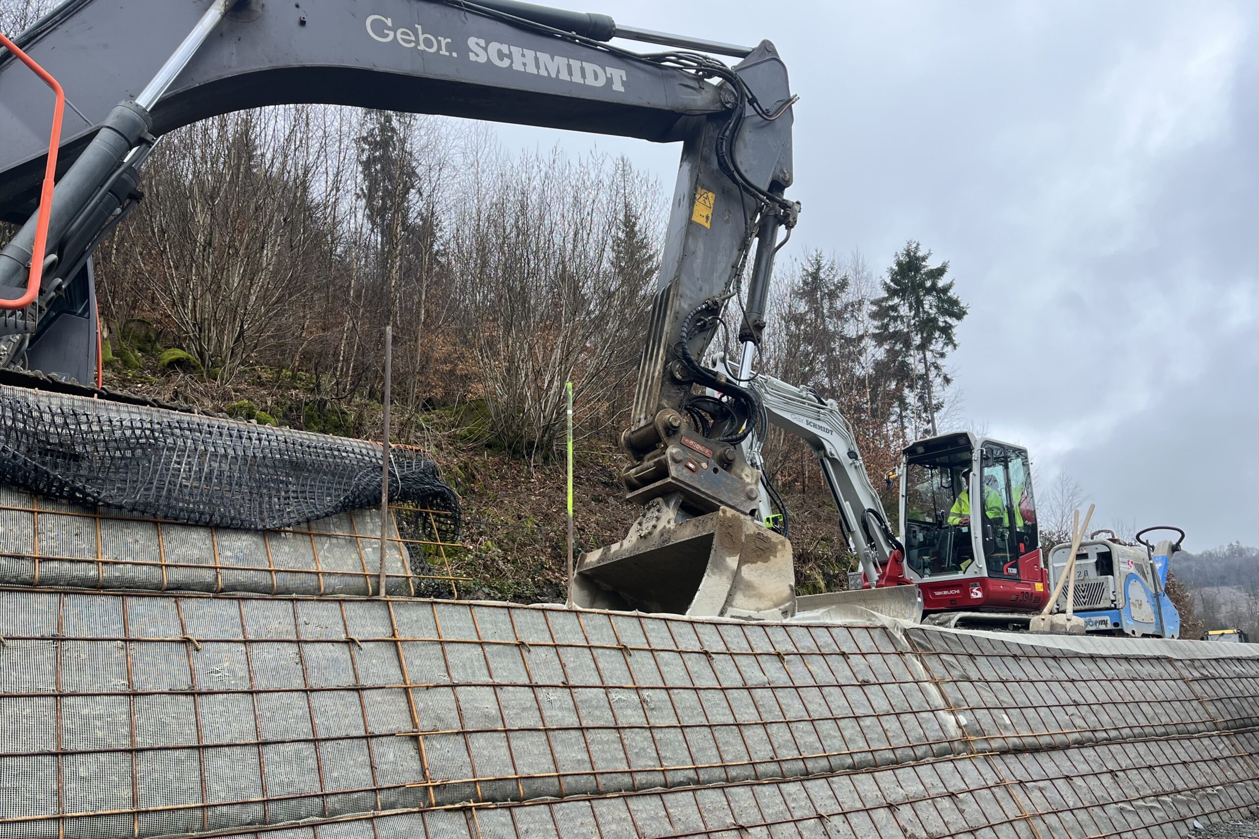 Bagger arbeitet an einer Stützmauer aus Beton und Kunststoffmatten an einer Straße. Im Hintergrund Bäume und ein weiterer Bagger.