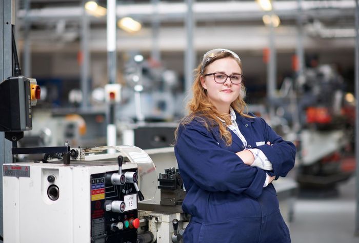Eine junge Frau mit roten Haaren und Brille steht verschränktarmig vor einer Drehmaschine in einer Werkstatt. Sie trägt einen blauen Arbeitskittel und eine Haube.