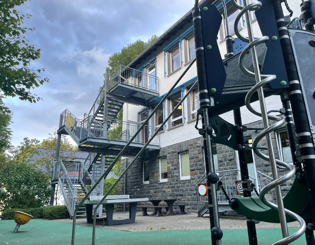 Ein mehrstöckiges Gebäude mit einer Metalltreppe im Freien. Im Vordergrund ein Spielplatz mit Klettergerüsten.