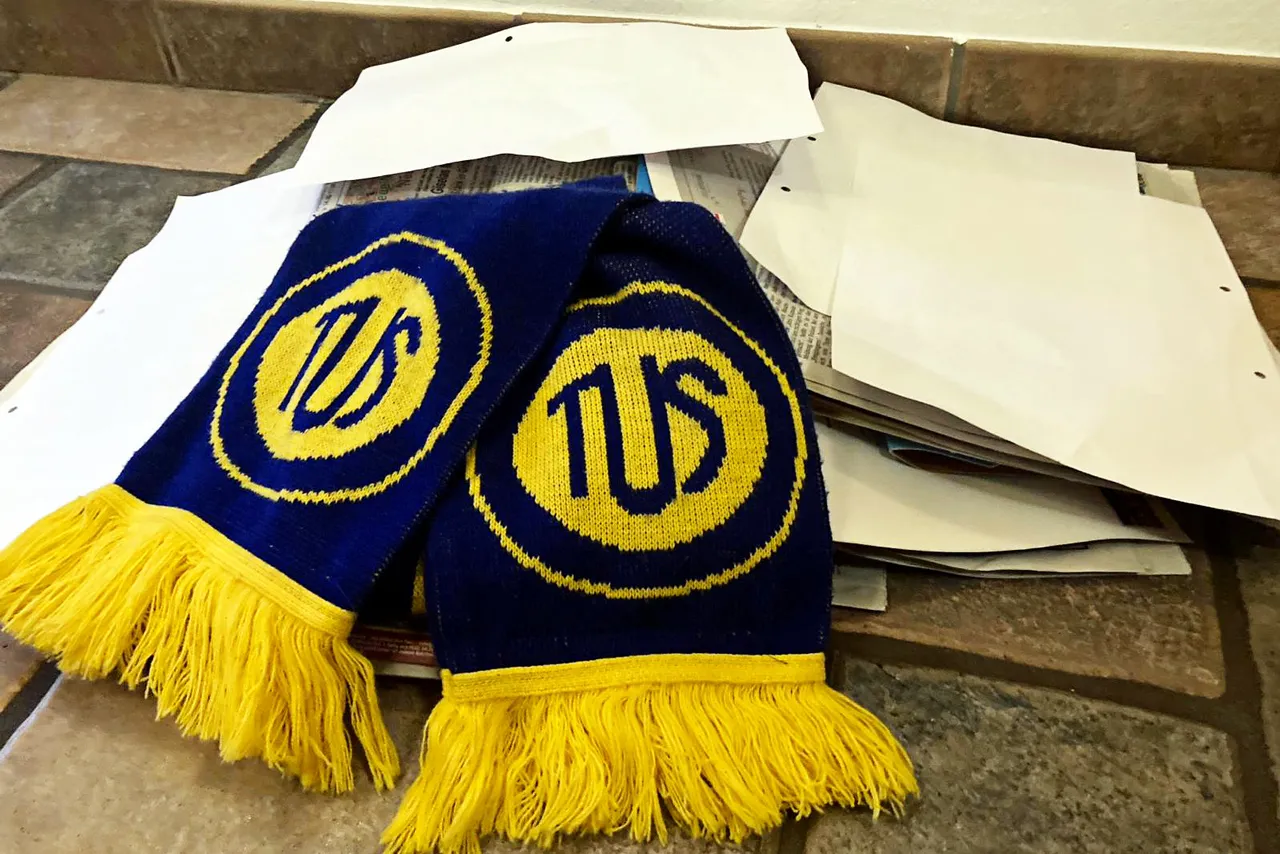 Zwei blaue TuS Plettenberg Schals mit gelben Fransen liegen auf einem Stapel Altpapier.