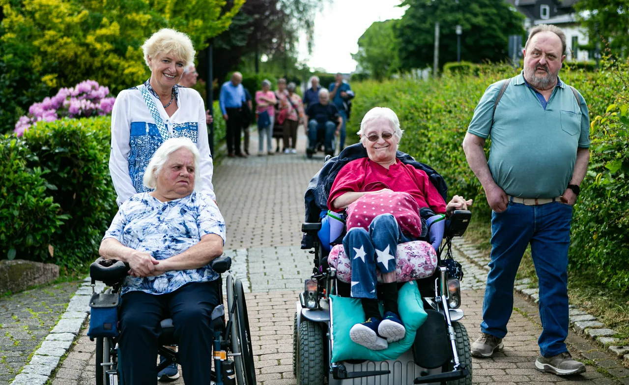 Zwei ältere Frauen im Rollstuhl und zwei weitere Personen gehen auf einem gepflasterten Weg, umgeben von Grün und einer Gruppe weiterer Menschen im Hintergrund.