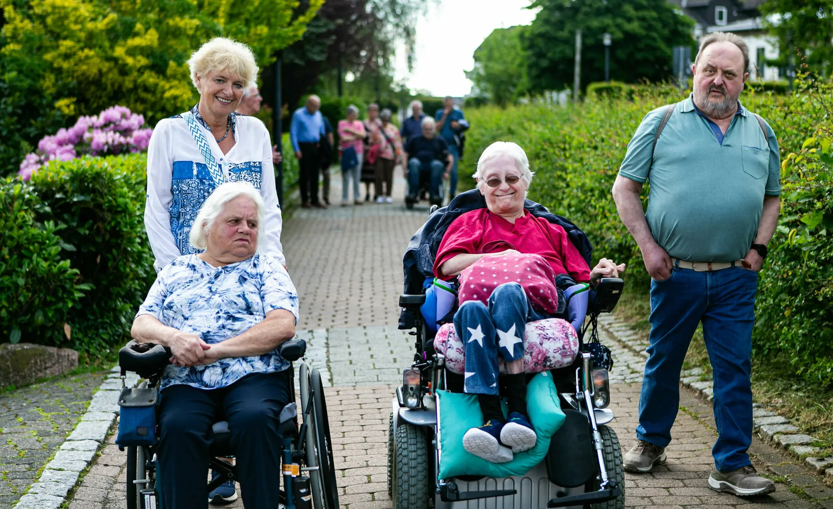 Zwei ältere Frauen im Rollstuhl und zwei weitere Personen gehen auf einem gepflasterten Weg, umgeben von Grün und einer Gruppe weiterer Menschen im Hintergrund.