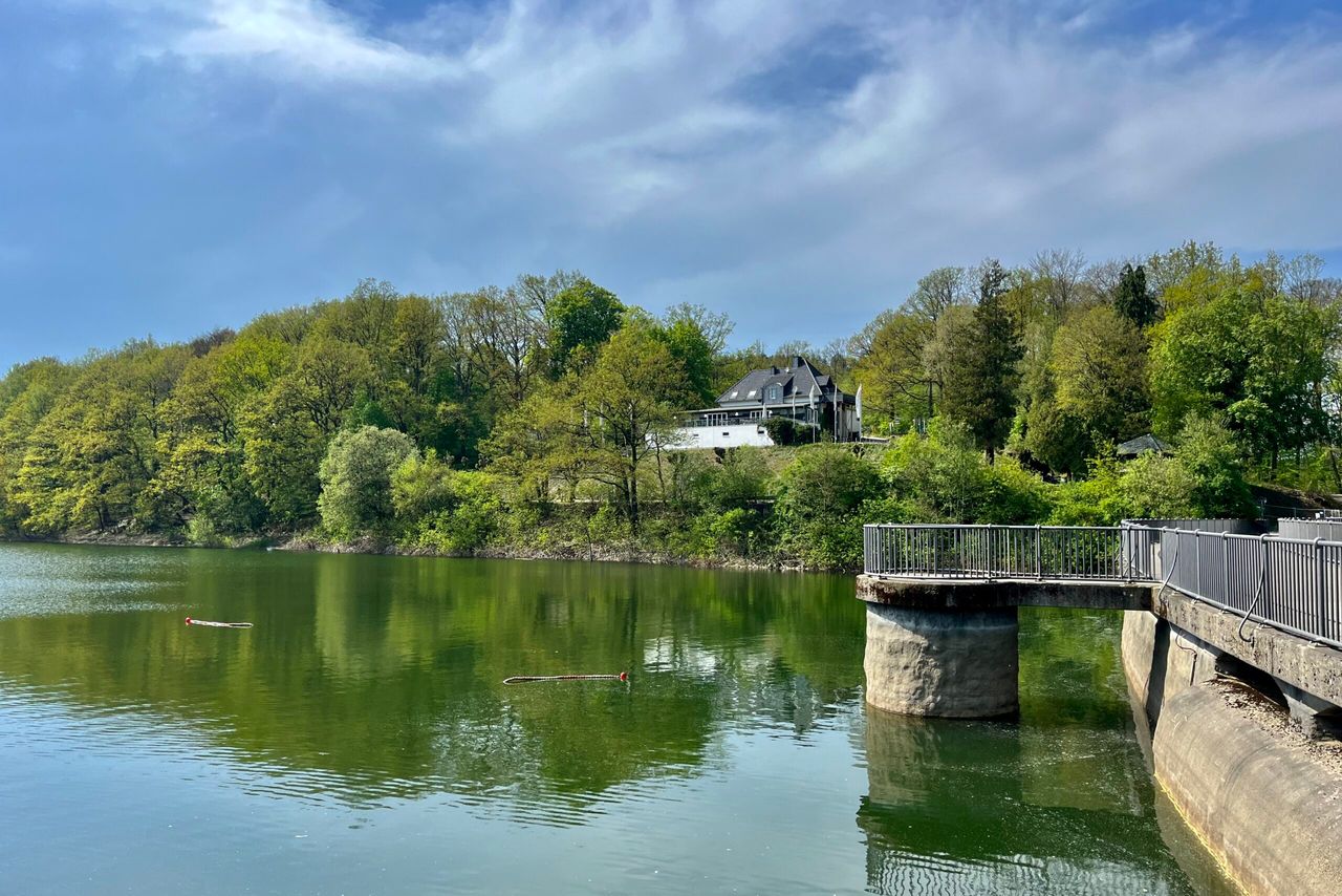 Blick auf die Glörtalsperre mit einem Gebäude auf einem bewaldeten Hügel und einer Brücke im Vordergrund.