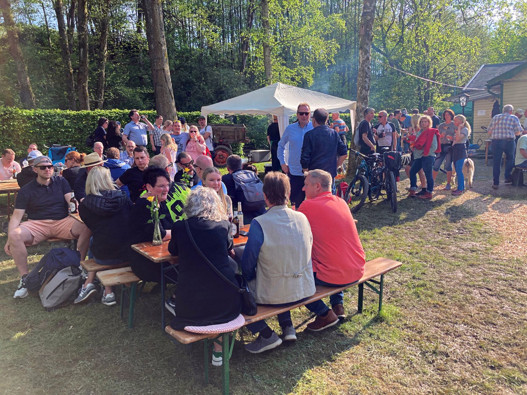 Menschen sitzen und stehen an Biertischen und auf einer Wiese zusammen, einige unter einem Zelt. Im Hintergrund Bäume und ein Gebäude.