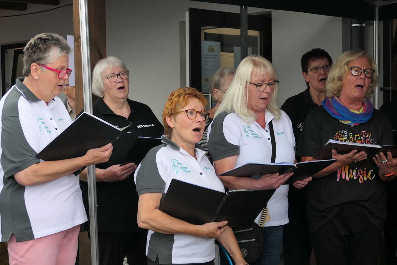 Ein gemischter Chor singt im Freien. Die Chormitglieder, hauptsächlich ältere Frauen, halten Notenblätter und singen mit geöffneten Mündern. Einige tragen T-Shirts mit dem Aufdruck 