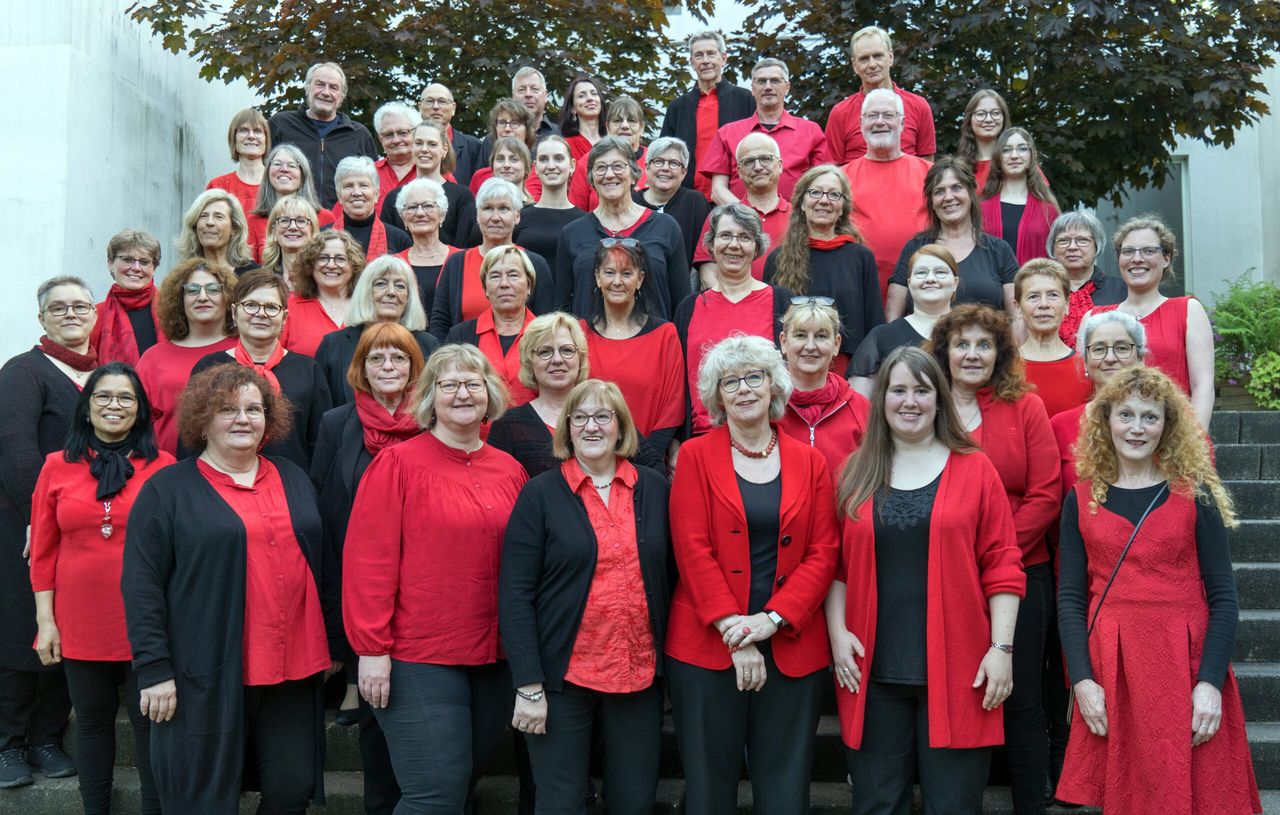 Eine große Gruppe von Menschen, hauptsächlich Frauen, posiert auf einer Treppe. Viele tragen rote Kleidung, einige schwarze. Das Bild zeigt den Gospelchor „Risecorn“, der zu einem Konzert einlädt.